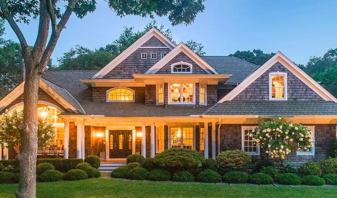 Best Airbnbs in the Hamptons - A large colonial style home in the evening with its lights on. It has a big front yard and is surrounded by a couple of trees.