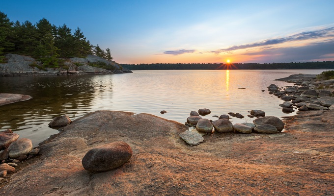 Places Like the Hamptons - A sunset visible from the rocky shore of a lake in Muskoka. It's surrounded by some trees.
