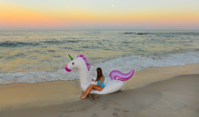 Places Like the Hamptons - A young woman sits on a unicorn pool float that's sitting on the shore of the ocean. It's just before sunset.