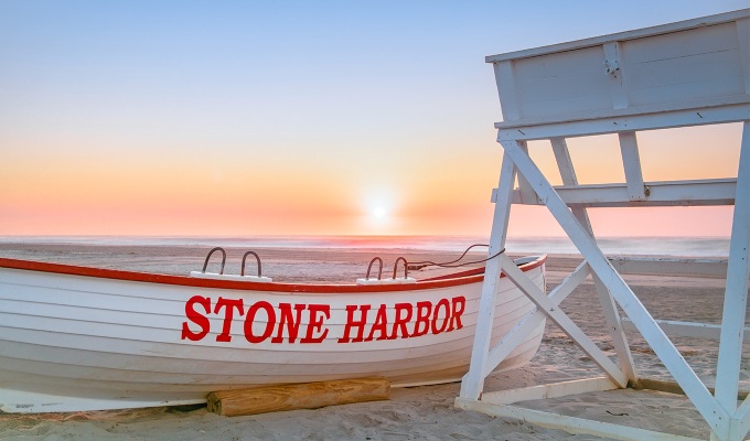 Places Like the Hamptons - A small boat with the words Stone Harbor on the side next to a lifeguard post. They are both on the shore of the beach at sunrise.