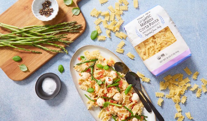 new at trader joe's june 2023: mafalda corta pasta on a serving platter, next to a bag of the dried pasta and a cutting board with asparagus